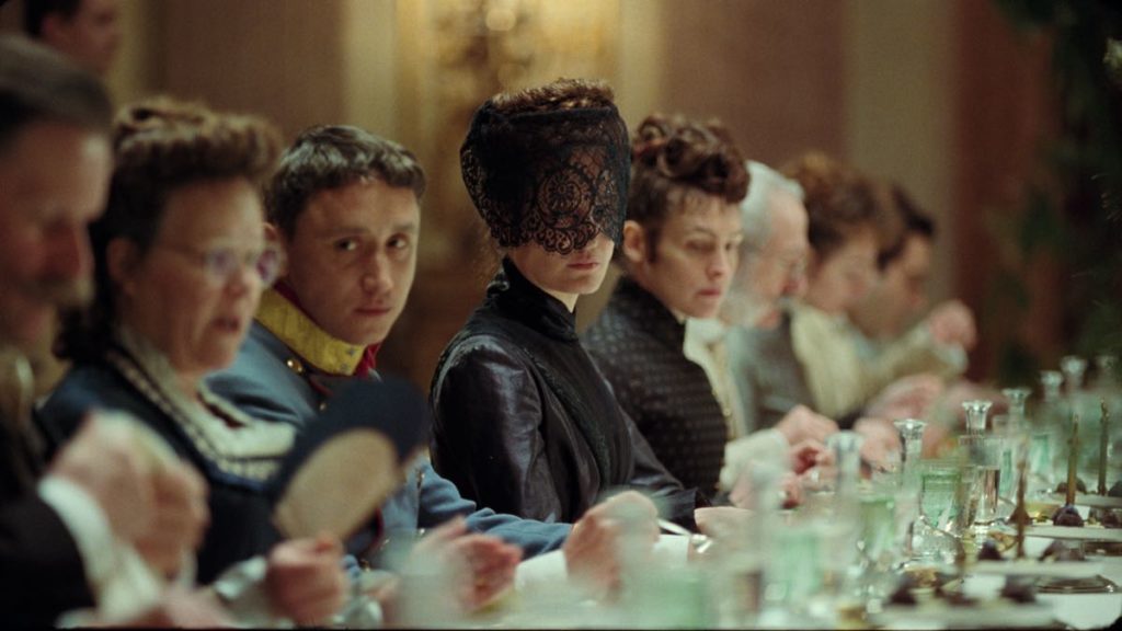 Empress Elisabeth (centre, veiled, played by Vicky Krieps) attends a formal state dinner in misery in Marie Kreutzer's film Corsage. She is seated next to her son (left) and her lady in waiting (right). Elisabeth is dressed in black and wearing black veil.