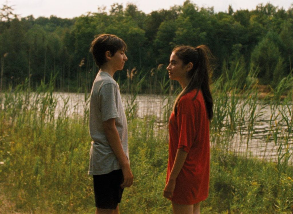 Still from Falcon Lake directed by Charlotte Lebon. Bastien (Joseph Engel, left) and Chloé (Sara Montpetit, right) stand close to one another at the side the lake amidst the tall grasses. Chloe is wearing an oversized red t-shirt, her long hair in a ponytail. Bastien is in a grey tshit and black swim trunks.