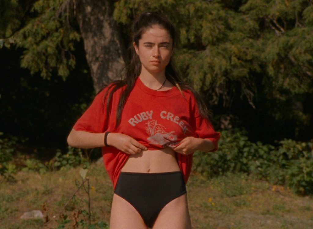 Chloé (Sara Montpetit) stands on the beach and begins to roll her red t-shirt up to reveal her chest to Bastien in Falcon Lake. Chloé is wearing black bikini bottoms and an oversized red t-shirt. She has rolled her t-shirt up above her midriff.