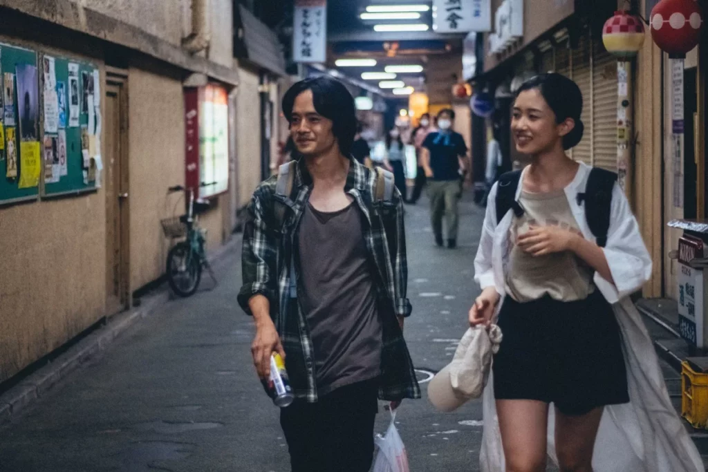 Teruo and Yo walk through a market at night, sharing a drink after their meet cute in Daigo Matsui's film Just Remembering