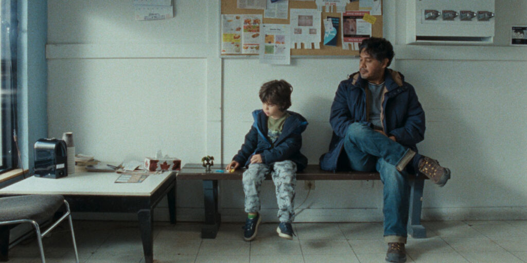 Still from Katherine Jerkovic's Coyote, one of the best films of TIFF 2022. A middle-aged man sits on a bench next to his grandson, who looks away from him in a school.