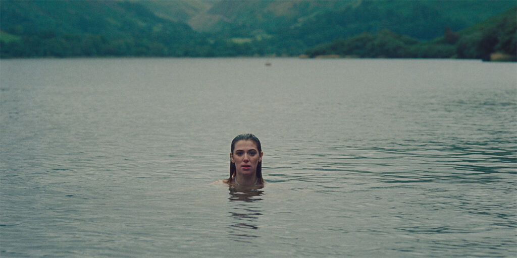 Honor Swinton Byrne stars in She Always Wins, one of the best and must-see shorts at TIFF 2022. In a wide shot, a young woman treads water in a lake, seen from the neck up, against a backdrop of green and blue rolling hills.