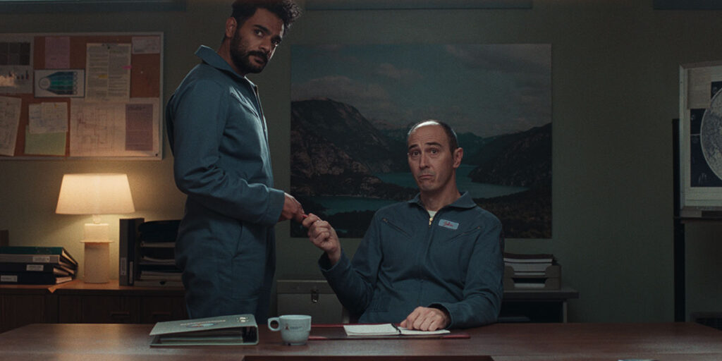 Hamza Haq (left) and Steve Laplante (right) in the film Viking, directed by Stéphane Lafleur, who is interviewed in this piece. Photo courtesy of TIFF.  Two men dressed in the same blue-grey space jumpsuit at a meeting table. One man is standing, and the other is seated, while both look into camera. They look like they're having a tug of war over a pen.