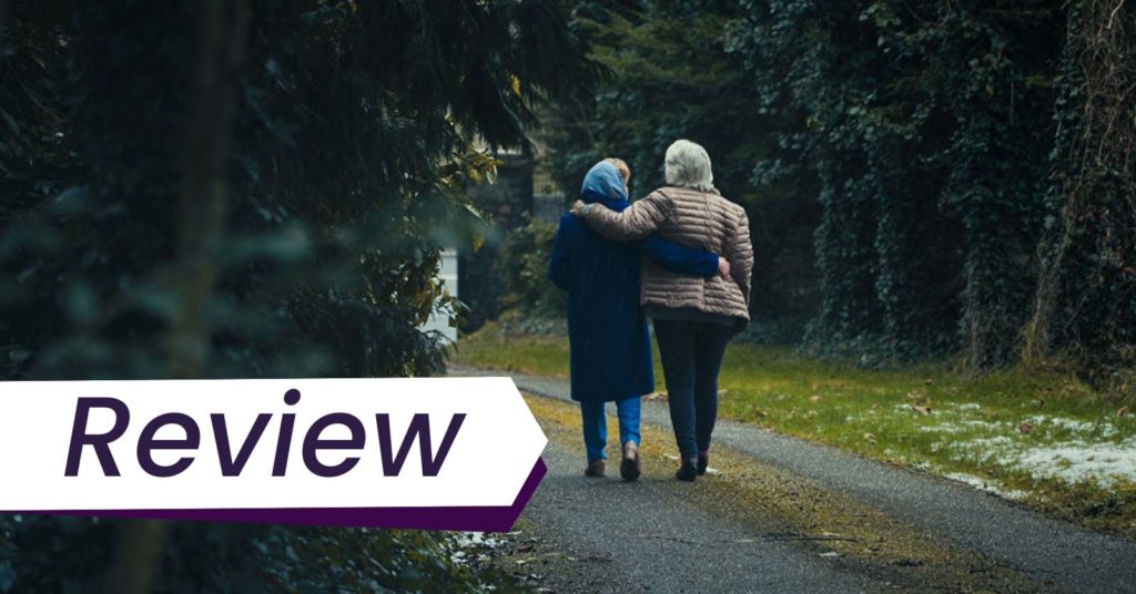 Still from Sinéad O’Shea’s film Pray for Our Sinners, which Alex Heeney reviews. Two elderly women featured in the documentary walk down a forested path, each with an arm around the other, seen from behind.