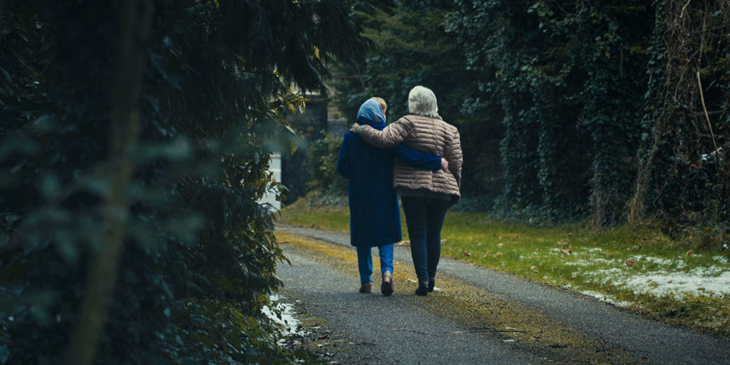 Still from Pray for Our Sinners, one of the best films of TIFF 2022. Two elderly women walk down a forested garden path arm in arm
