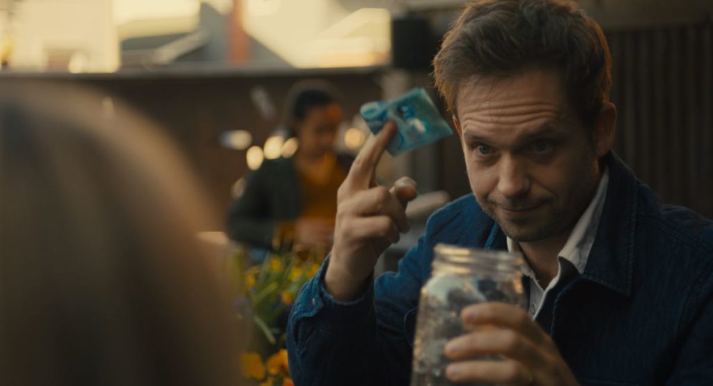 Patrick J. Adams stars in Lindsay MacKay's film, The Swearing Jar, which Alex Heeney reviews. Simon holds up a blue Canadian five-dollar bill above the titular swearing jar. The back of Carey's head can be seen in the front left of the frame.