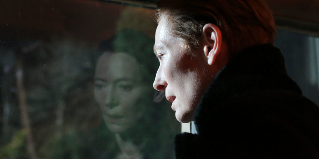 Tilda Swinton stars as Julie in Joanna Hogg's The Eternal Daughter, one of the best films at TIFF 2022. A middle-aged woman in a turtleneck and short hair looks out the window at the forest and her reflection is seen in the window.