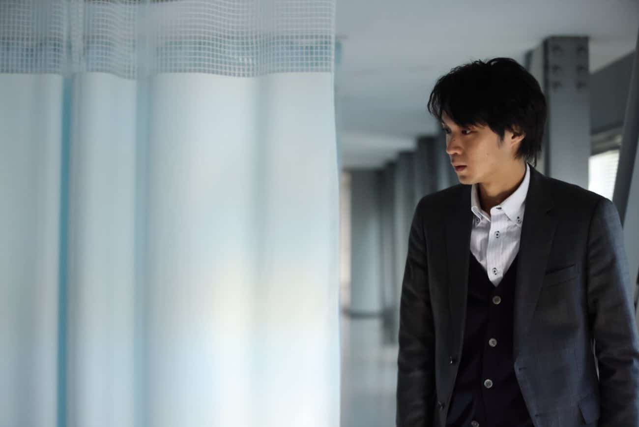 Hiromu in a grey suit looks at his (offscreen) dead uncle, while standing next to a blue hospital curtain in the Plan 75 centre. In this interview, Chie Hayakawa talks about getting actor Isomura Hayato to underplay the scene.