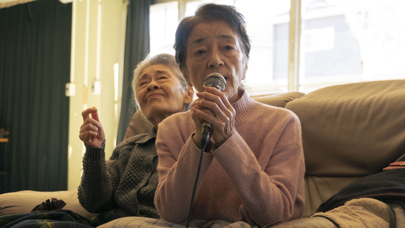 Michi (Chieko Baisho) sings karaoke with her friends in Chie Hayakawa's Plan 75. Michi is a 78-year-old woman with short grey hair wearing a pink turtleneck. She is seated next to her friend, an older woman whose eyes are closed and hand in the air, enjoying Michi's singing. They are both seated on a beige couch. In this interview, Chie Hayakawa discussed how women tend to have more community than men do in Japan.