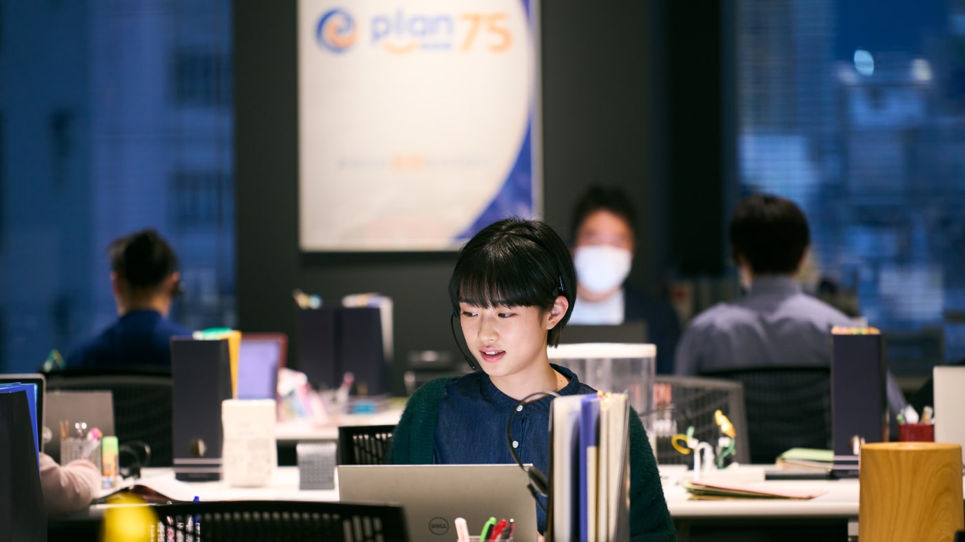 Yoko (a young woman with short hair in a forest green sweater) sits in front of a laptop with a headset on, in front of the Plan 75 sign in her office, with her coworkers behind her in Chie Hayakawa's Plan 75. In this interview, Hayakawa talks about designing the office to be cold and grey and showing us what is happening behind Yoko in her world.