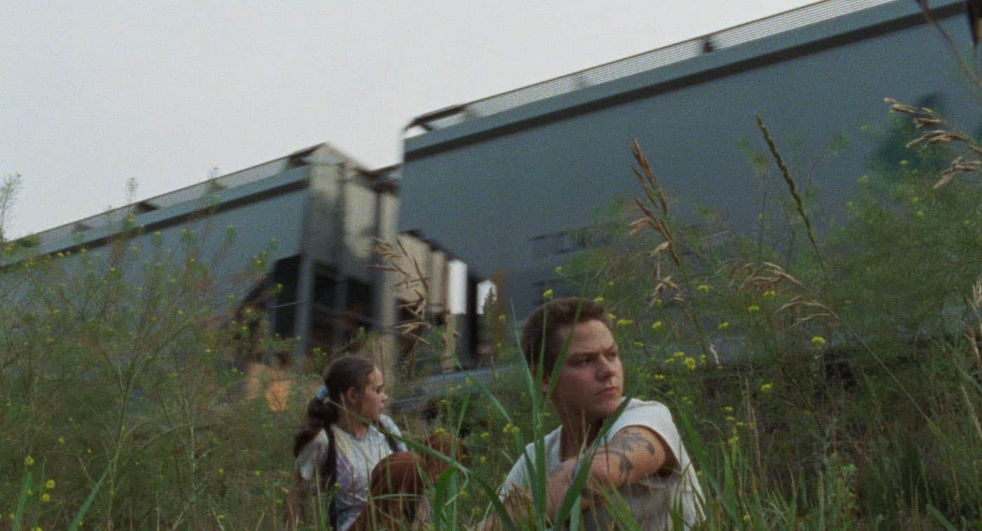 Still of Whitney (left) and Kyle (right) in The Maiden directed by Graham Foy. A teenage girl and boy dressed in white t-shirts sit in the grass in front of a passing cargo train.