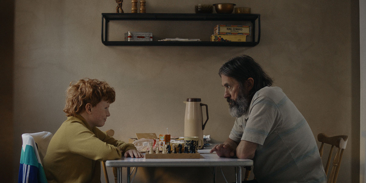 A red-headed boy and a grey-bearded middle-aged man play chess at the kitchen table in Solitude.