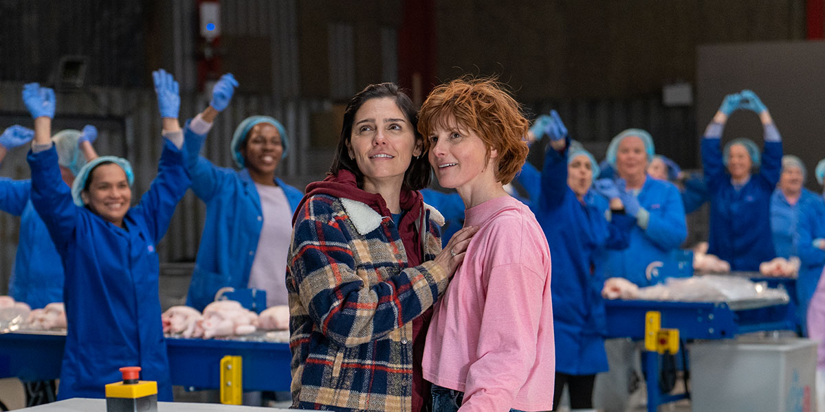 Two women embrace in a chicken factory, surrounding by cheering employees in blue jumpsuits, gloves, and hairnets in Janis Pugh's Chuck Chuck Baby.