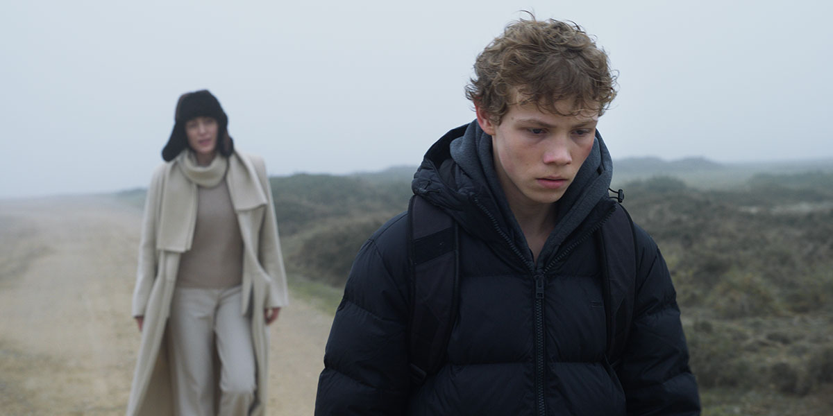 A middle-aged woman in a brown winter hat, white ankle-length wool coat, white sweater and pants, follows behind a gloomy-looking teenage boy dressed in dark colours, across a rocky, foggy landscape in Hanna Slak's film Not a Word.