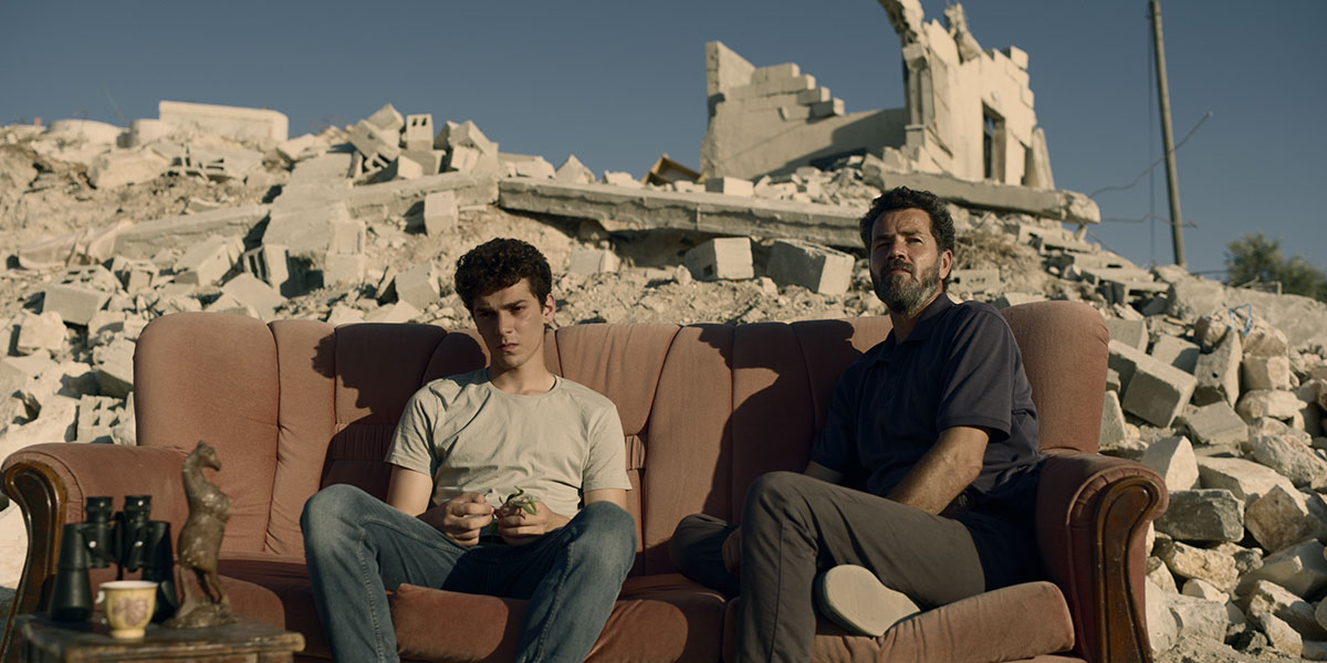 A teenage boy and his teacher sit on a red couch against the backdrop of the boy's recently demolished house in The Teacher, directed by Farah Nabulsi.