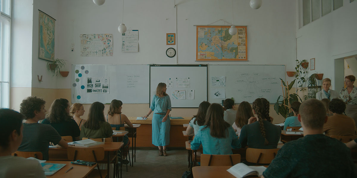 A teacher dressed in blue, stands in front of a class of teenagers, and looks at the door where other teachers have arrived to supervise her in Without Air, one of the best acquisition titles at TIFF 2023.