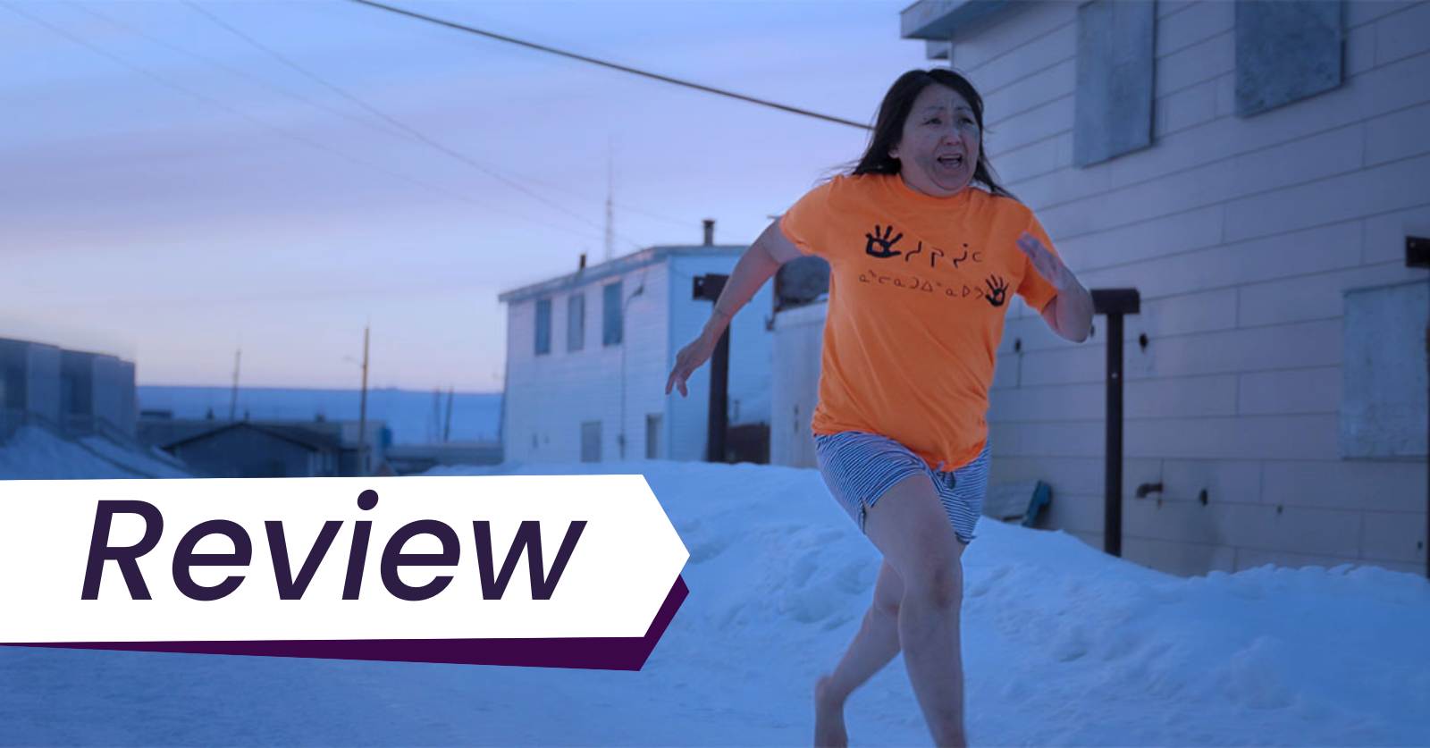 An Inuk woman (Lucy Tulugarjuk) in an orange tshirt and grey shorts runs through the snow barefoot at dusk in Tautuktavuk (What We See).