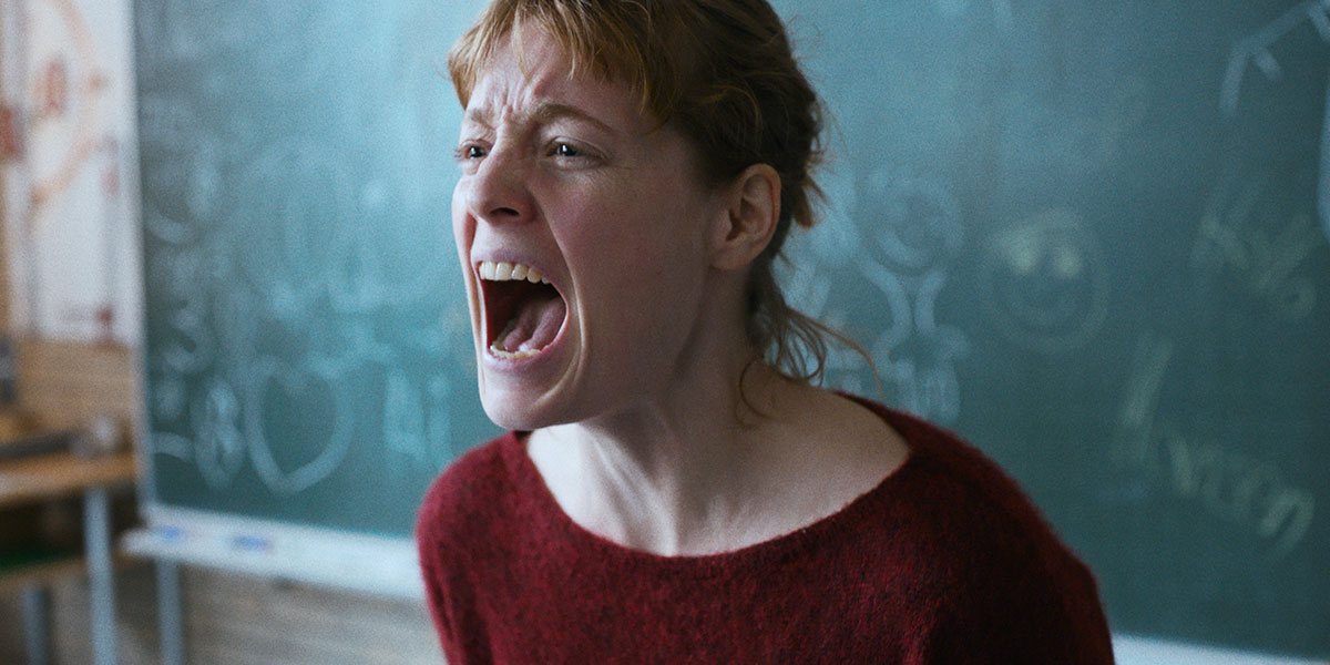 A woman with red hair, pulled back in a ponytail, wearing a red sweater, screams while standing in front of a blackboard in her classroom in Ilker Çatak's The Teachers' Lounge