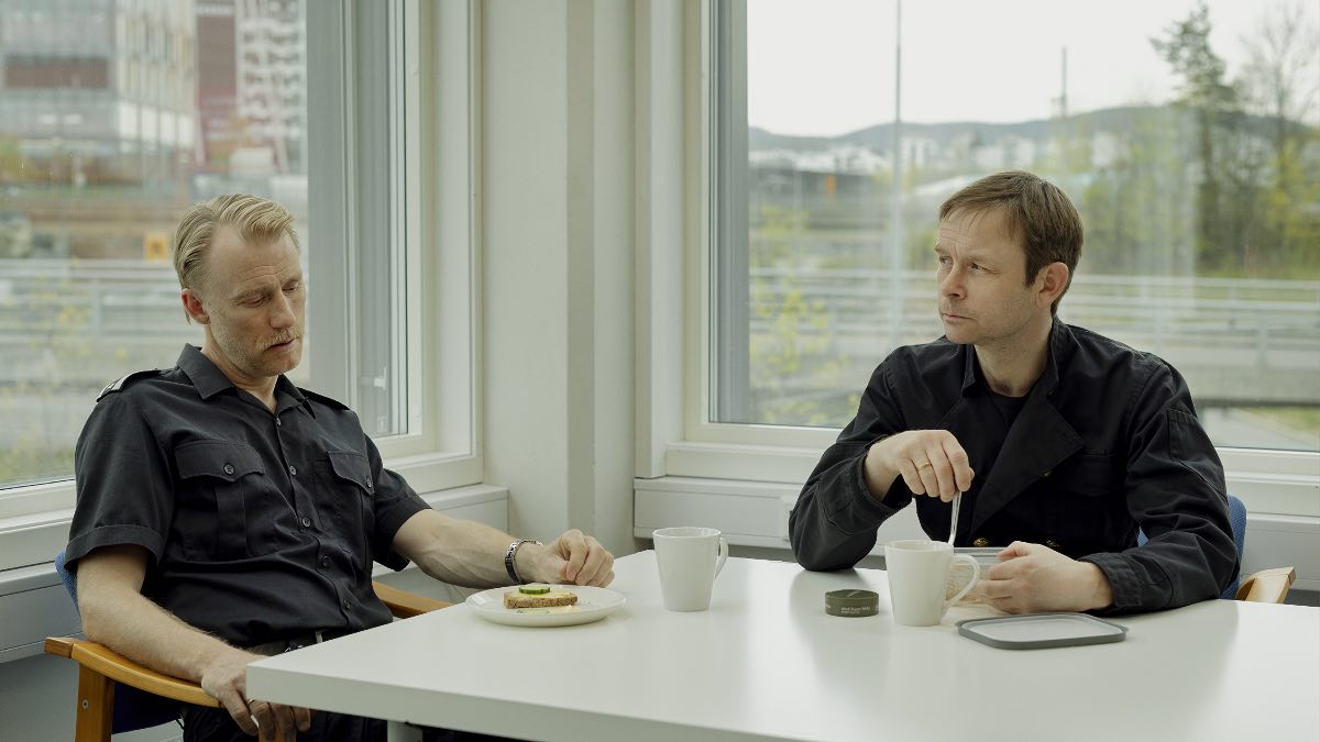 Thorbjørn (left) reveals his dream to Jan (right) who reveals he just had sex with a man in an early scene of Dag Johan Haugerud's film Sex at the Berlinale. Two white men dressed in black sit at a white table at right angles in the corner of a room with windows behind them.