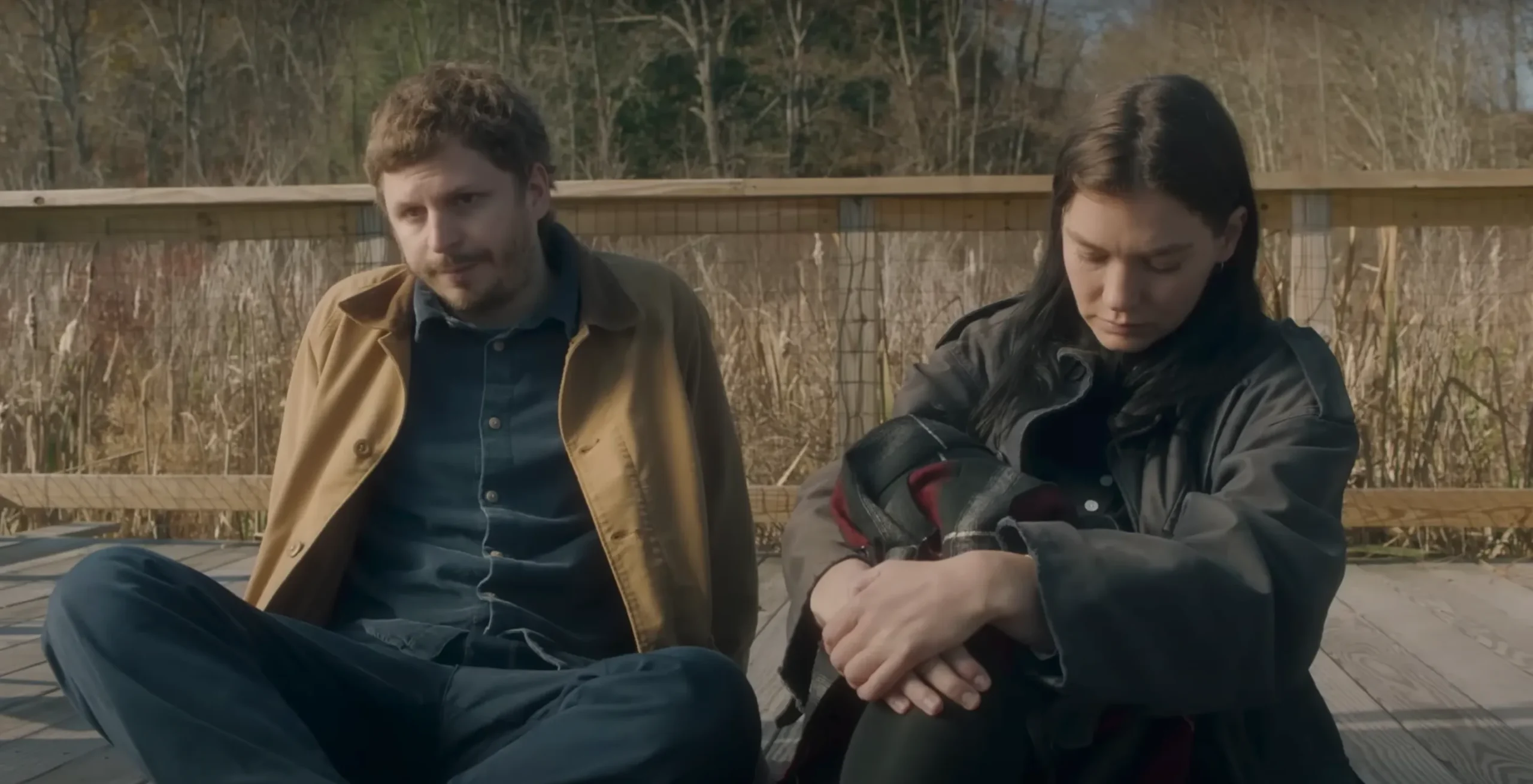 Michael Cera and Hannah Gross star in The Adults by Dustin Guy Defa. A blond man in a tan workcoat (left), a denim shirt, and blue slacks and a woman dressed all in black with dark hair sit next to each other on a boardwalk, avoiding eye contact. 