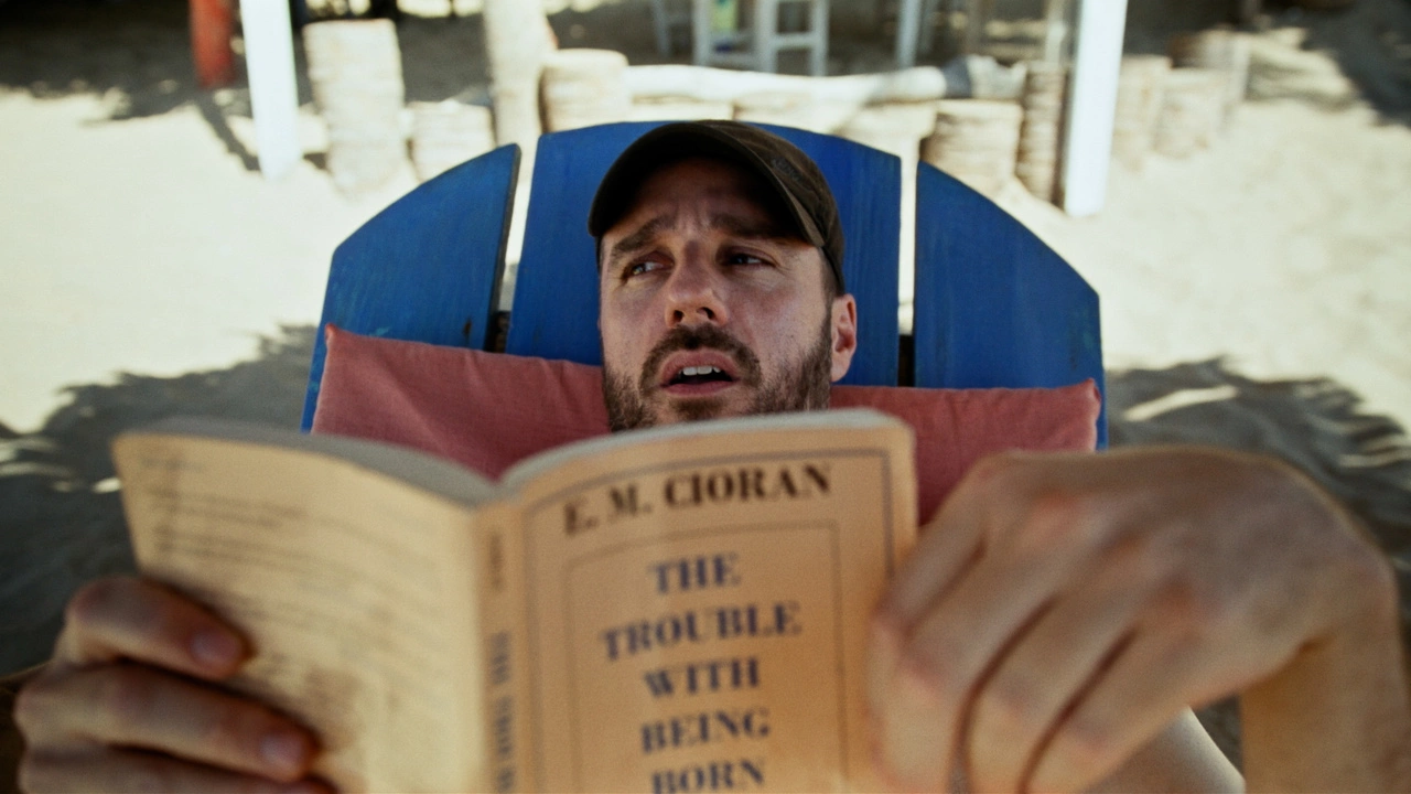 Sebastián Silva stars in Rotting in the Sun, one of the best films of 2023. A Latinx man in a black baseball cap lies on a a beach in a blue chair reading The Trouble with Being Born.