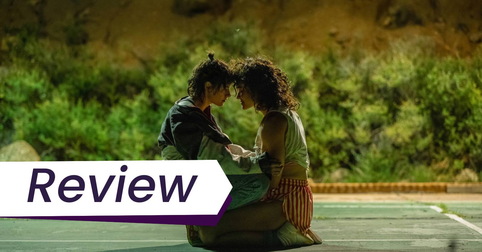 Still of  Lou (Kristen Stewart) and Jackie (Katy O'Brian) in Rose Glass's film Love Lies Bleeding, which Lena Wilson Reviews. Photo courtesy of A24 Films. Two women sit kneeling as if in an embrace on a tennis court, their heads together.