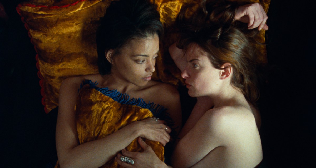 Still from When Night is Falling, which Patricia Rozema discusses in this interview. A Black woman and a white woman lie in bed on golden pillows and blankets, looking at each other.