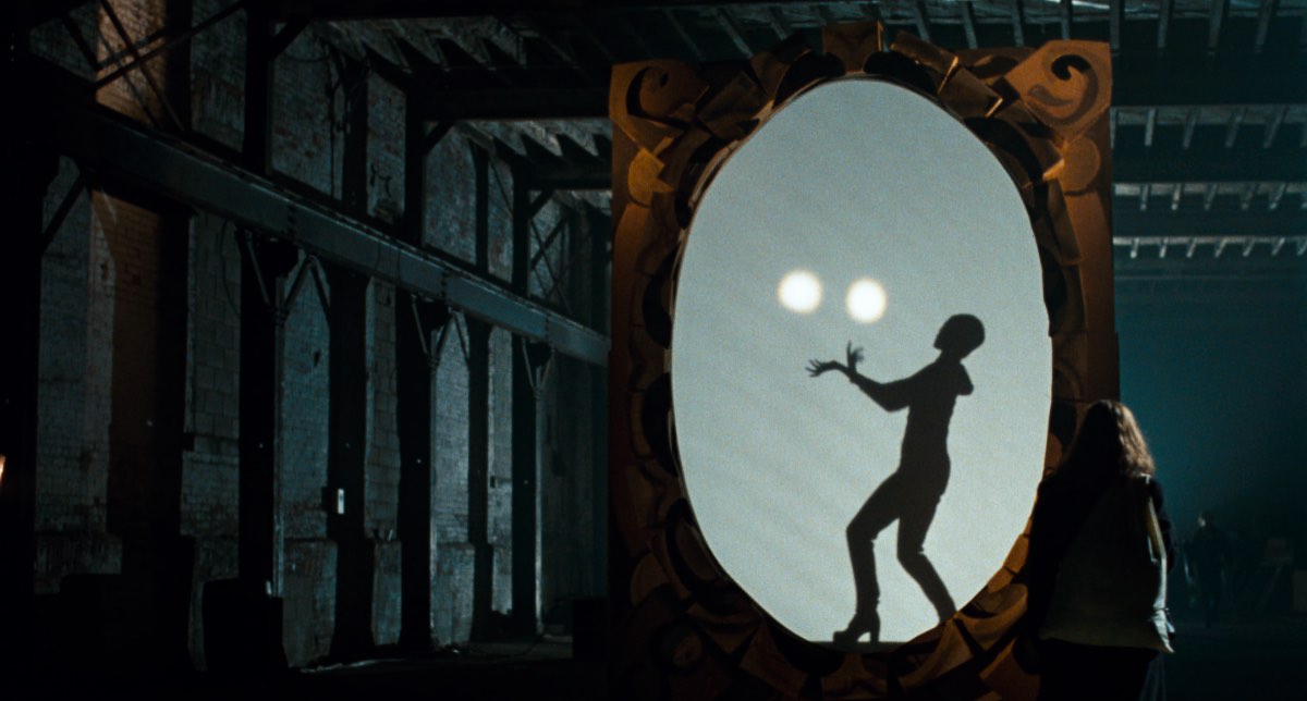 Still from When Night is Falling of the circus, which Patricia Rozema discusses in this interview. Image courtesy of Kino Lorber. A woman juggling as seen through a large picture frame and screen inside a warehouse. Another woman is watching, back turned to the camera.