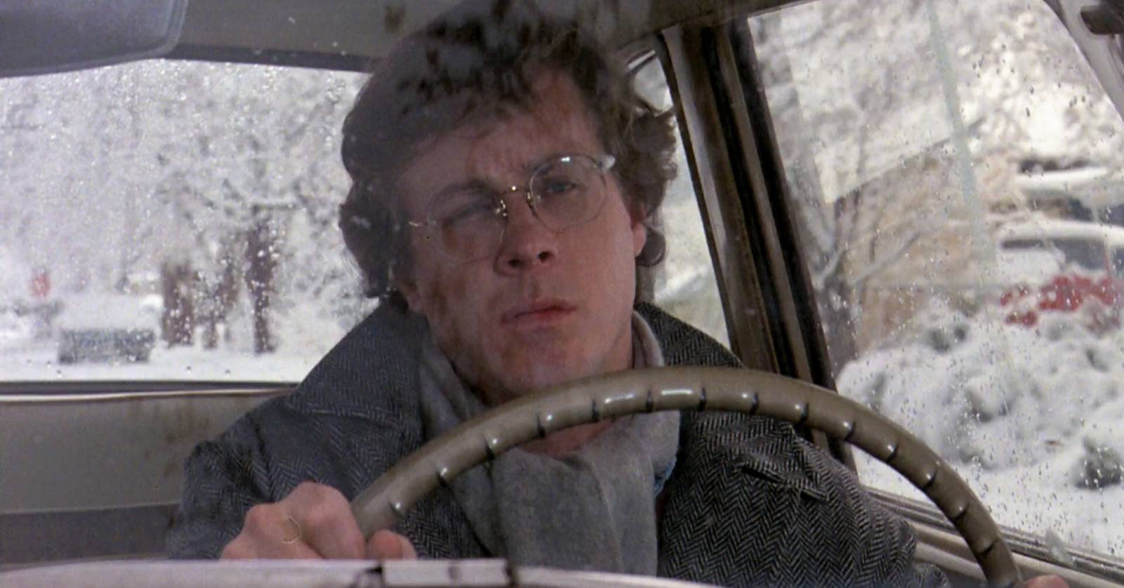 John Heard as Charles in Chilly Scenes of Winter, now in the Criterion Collection, which we review. A man with glasses in a grey overcoat drives in the Utah winter