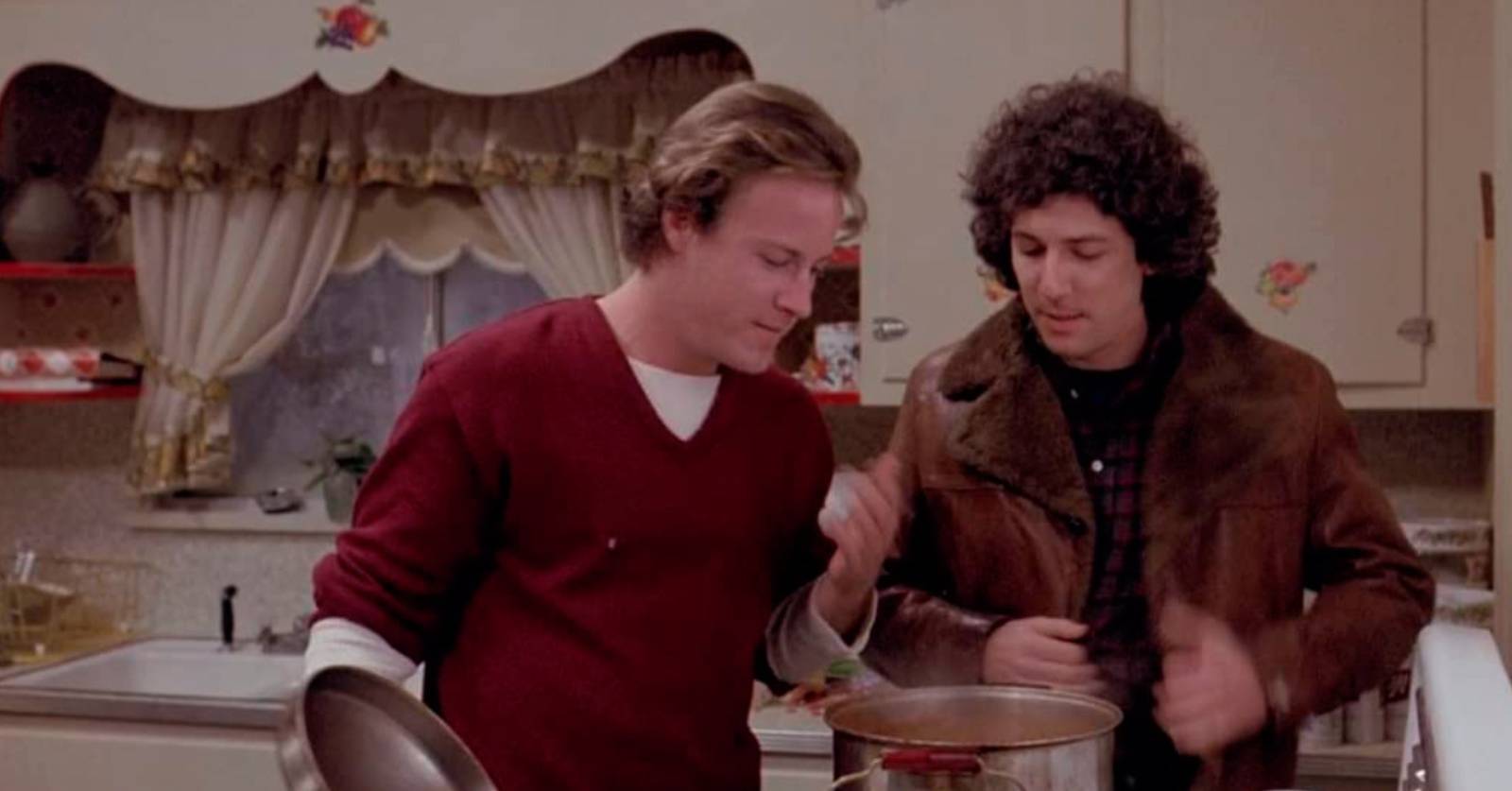 Two men in a kitchen look inside a pot of chili .