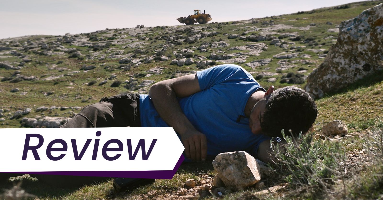 Still from the film No Other Land, one of the highlights of the Visions du Réel Film Festival. A man in a blue t-shirt lies on the ground with a tank driving by in the background.