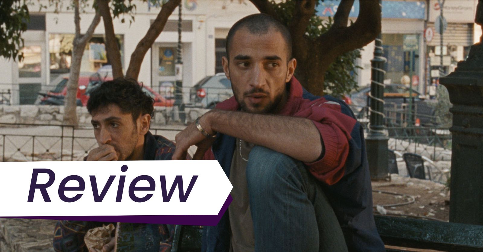 Mahmood Bakri stars in Mahdi Fleifel's  film To a Land Unknown, which has its world premiere at TIFF 2024. Photo Courtesy of TIFF. Two Palestinian man sit on a park bench in Athens, one of them is wearing a red shirt and jeans and has a buzz cut, and the other has short hair and sideburns and eating something.