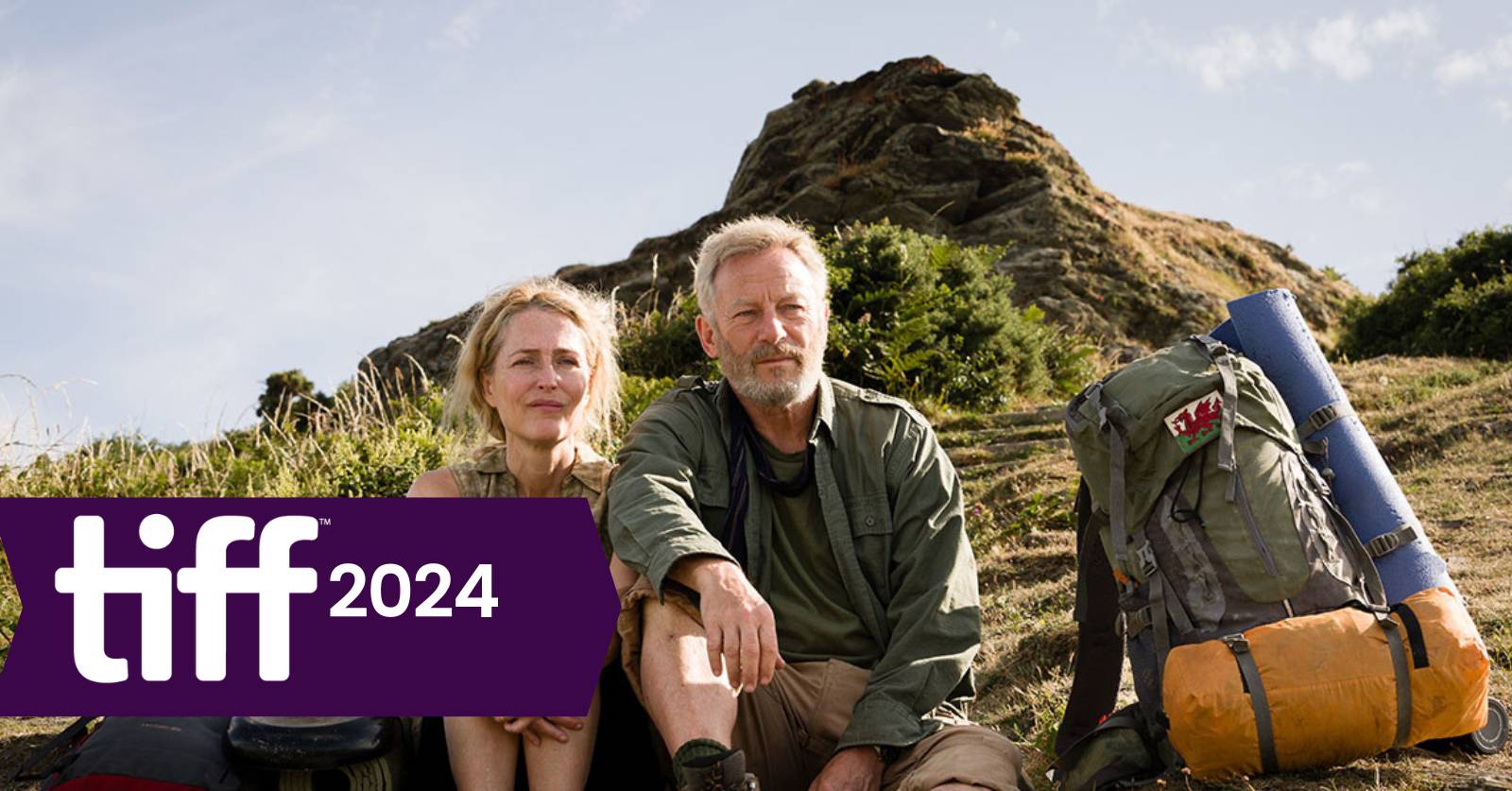 Still of Gillian Anderson (left) and Jason Isaacs (right) in Marianne Elliott's film adaptation of The Salt Path.  Alex interviews Marianne Elliott about The Salt Path on the podcast today. A middle-aged white woman and white man with hiking gear sit side by side on a hill.