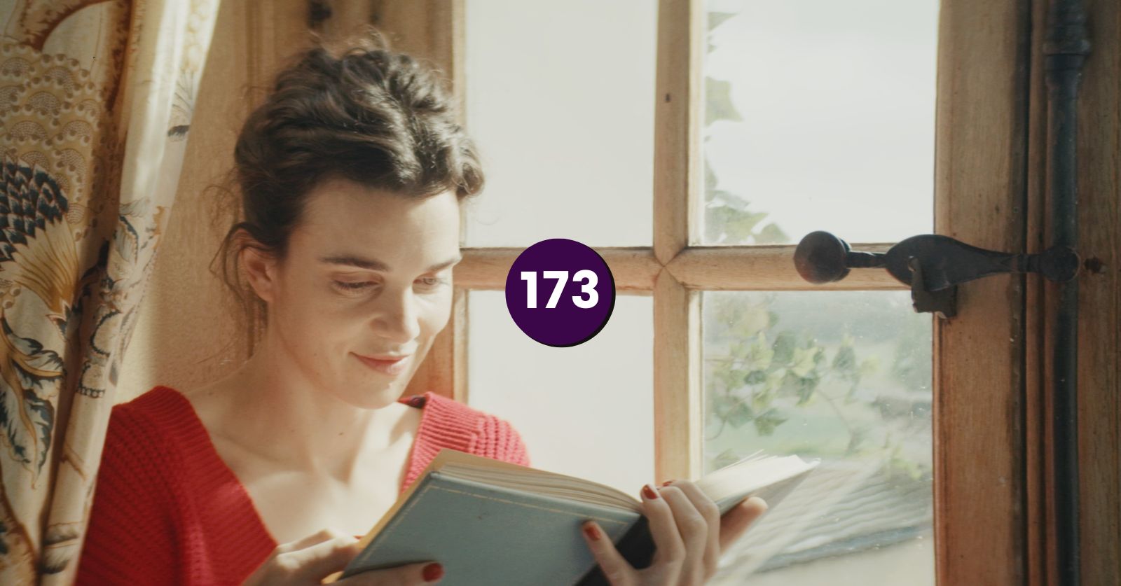 We did into the beginning and ending of Jane Austen Wrecked My Life on the podcast, but since we don't have a still from there, here's a still in the right spirit from the film. A woman with brown hair in a red sweater reads a book next to a window and curtain.